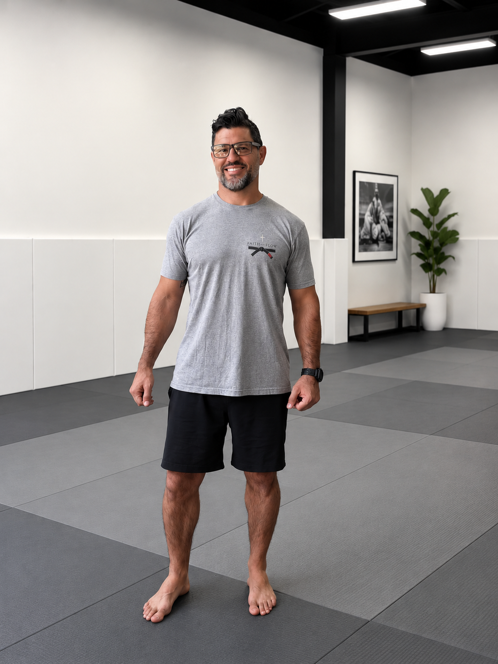 A man wearing a heather gray athletic t-shirt with a small black logo on the chest and black shorts, standing barefoot on gray mats inside a modern fitness studio.