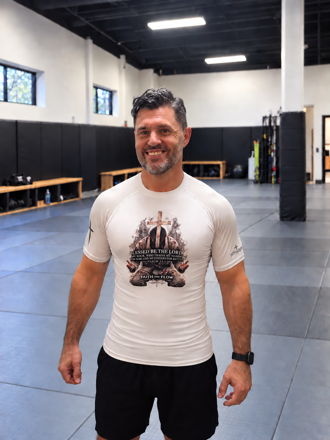 A smiling man in a gym wearing a white short-sleeve athletic t-shirt with a detailed graphic print on the front, paired with black shorts.
